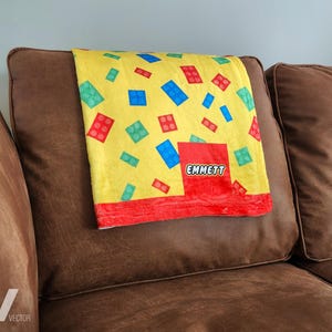 May include: A yellow blanket with a colorful brick pattern, featuring red, blue, and green blocks. The blanket has a red border and a red square with the name "EMMETT" in white. The blanket is draped over a brown couch cushion.