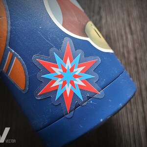 May include: Close-up of a blue water bottle with a starburst sticker in red, white, and blue. The bottle also features orange and white graphics. The sticker is transparent and has a star shape.