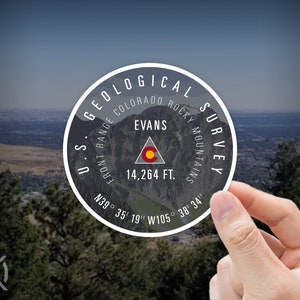 May include: A round sticker with a white border and black text. The text reads "U.S. Geological Survey, Colorado Rocky Mountains, Front Range, Evans, 14,264 ft., N39° 35' 19" W105° 38' 34"". The sticker features a stylized mountain range and a red and yellow triangle with a white outline.