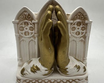 May include: A ceramic bookend depicting hands in prayer. The hands are gold, and the base and architectural features are white with gold detailing. The design incorporates gothic arches and circular patterns, creating a religious theme.