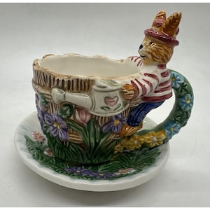 May include: A whimsical ceramic teacup and saucer set. The teacup is shaped like a basket with a bunny figure on the side. The handle is adorned with blue and yellow flowers. The saucer and cup are decorated with flowers and greenery.