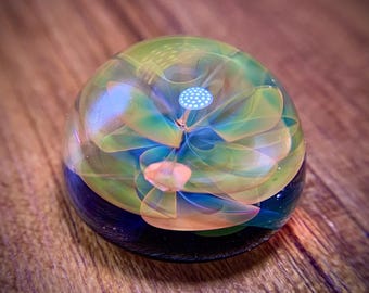 May include: A clear glass paperweight with a floral design. The interior features a layered, translucent flower in shades of green, yellow, and blue, with a small, white, dotted center. The base is a dark blue and black. The paperweight sits on a wooden surface.