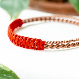 May include: Copper bracelet with a twisted design and a section wrapped in bright orange thread. The bracelet is set against a white background with green foliage in the corners.