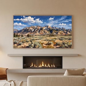 Op de afbeelding: Een rechthoekig landschapsschilderij met een bergketen met besneeuwde toppen onder een helderblauwe lucht met witte wolken, ingelijst in een dunne houten lijst boven een lineaire open haard in een woonkamer met neutrale tinten.