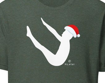 May include: Dark green t-shirt featuring a white graphic of a person doing Pilates, wearing a Santa hat. The shirt has the word "PILATAY" printed in small white letters. The shirt is made of a soft material.
