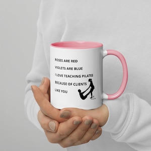 May include: White ceramic mug with a pink rim and handle. The mug has black text that reads "Roses are red, Violets are blue, I love teaching Pilates because of clients like you." There is a black silhouette of two people doing a Pilates exercise.