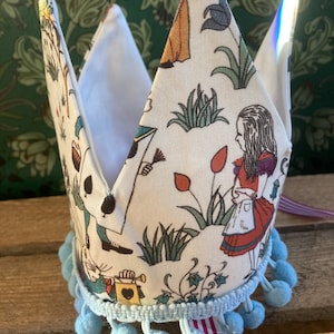May include: A white fabric crown with a blue pom-pom trim and a colorful Alice in Wonderland print. The crown features illustrations of Alice, the Mad Hatter, and other characters from the book.