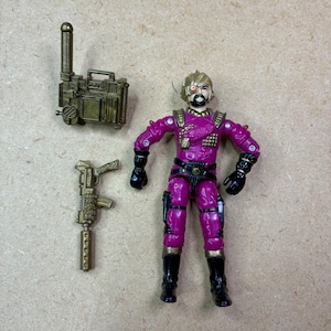 Peut inclure: Une figurine articulée violette avec des accents d'armure dorés, une barbe noire et un cache-œil, accompagnée d'un appareil de communication rectangulaire doré avec antenne et d'un fusil doré avec un long canon.