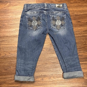 May include: Blue denim capri trousers featuring a faded wash and rolled cuffs. The back pockets are decorated with embroidered cross and rose motifs accented with small metallic studs. A rectangular brand label is stitched on the waistband.