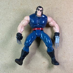 Könnte beinhalten: Eine muskulöse Actionfigur mit schwarzen Haaren, Koteletten und einer Augenklappe. Die Figur trägt einen dunkelblauen ärmellosen Anzug mit rotem Gürtel, schwarzen Handschuhen und schwarzen Stiefeln. Silberfarbene Krallen mit drei Klingen.
