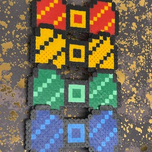 May include: Four colorful bow tie designs made from small, fused plastic beads. The bow ties are red, yellow, green, and blue, each with a black border and a square center. The background is a dark gray with gold splatter.