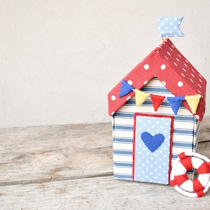 Op de afbeelding: Een rood met witte stippen strandhuisje met blauw-wit gestreepte muren en een blauwe deur met een hart. Het huisje heeft een rood, geel en blauwe vlaggenlijn en een blauw met witte stippen vlag bovenop. Er ligt een rood-witte reddingsboei voor het huisje.