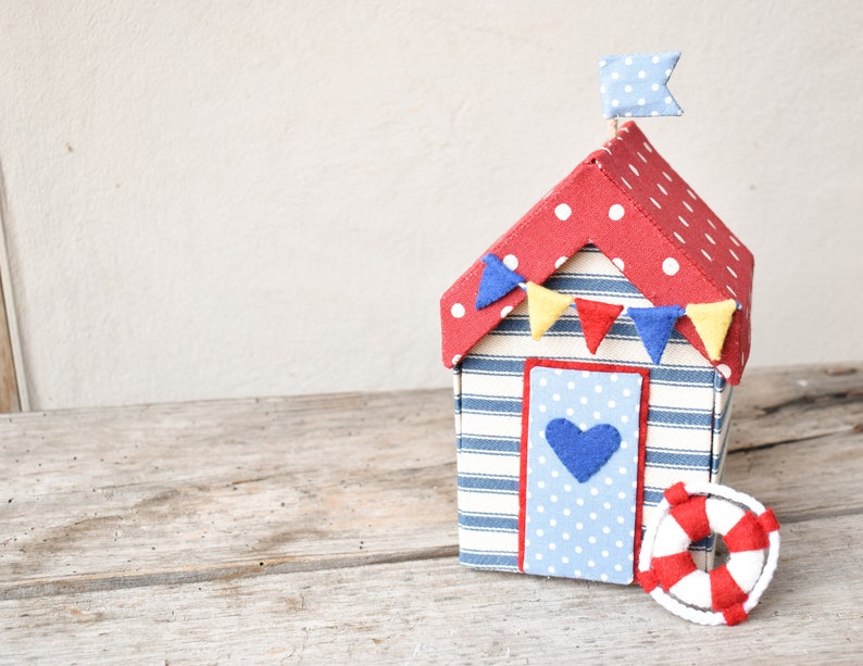 Op de afbeelding: Een rood met witte stippen strandhuisje met blauw-wit gestreepte muren en een blauwe deur met een hart. Het huisje heeft een rood, geel en blauwe vlaggenlijn en een blauw met witte stippen vlag bovenop. Er ligt een rood-witte reddingsboei voor het huisje.