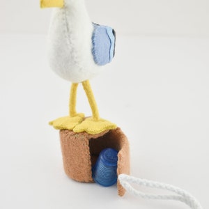 Op de afbeelding: Een witte viltmeeuw met gele poten en een blauwe vleugel staat op een bruine viltdoos met een wit koord. De doos is open en er zit een blauwe plastic vingerhoed in.