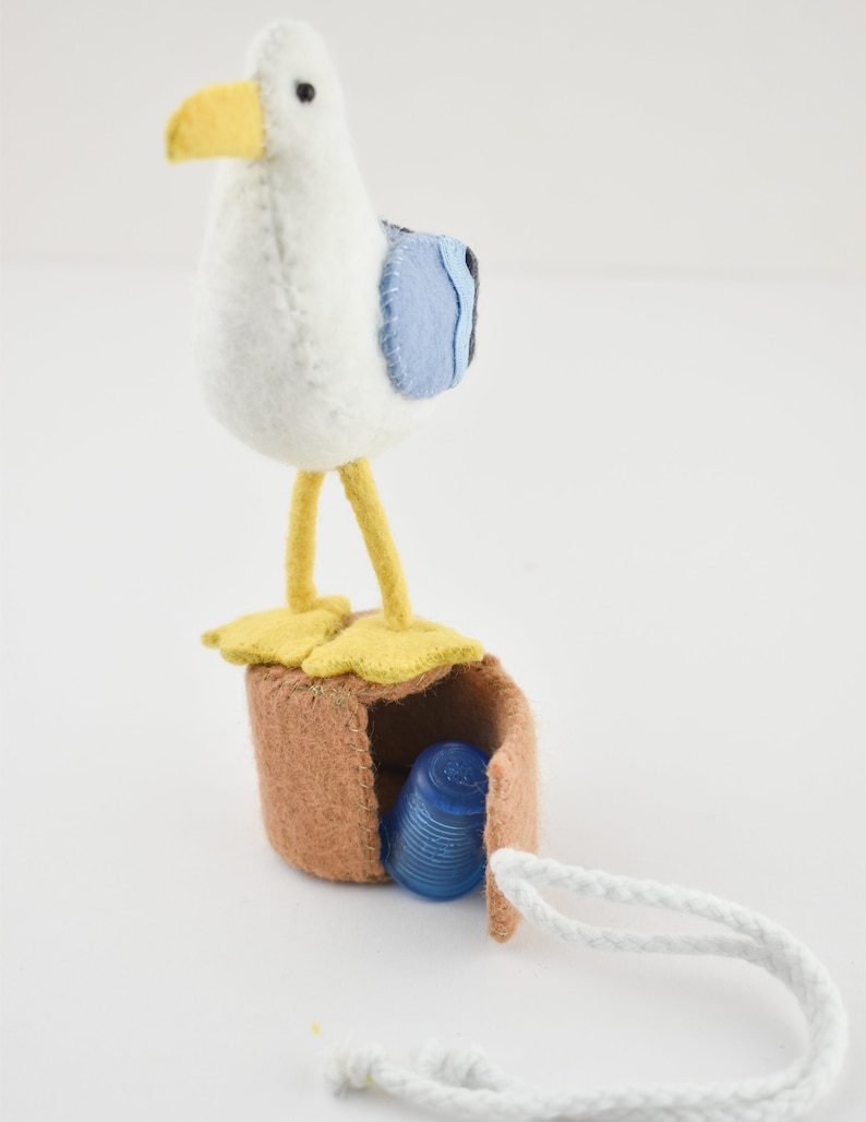 Op de afbeelding: Een witte viltmeeuw met gele poten en een blauwe vleugel staat op een bruine viltdoos met een wit koord. De doos is open en er zit een blauwe plastic vingerhoed in.