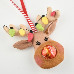 Puede incluir: Un adorno de reno de fieltro con un lazo de cinta rojo y blanco a rayas. El reno tiene una nariz roja con un caramelo de chocolate dentro, y sus cuernos est&aacute;n decorados con luces de fieltro verde, amarillo y rojo.