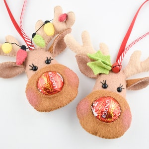 Puede incluir: Dos adornos de reno de fieltro con cintas de suspensi&oacute;n a rayas rojas y blancas. Cada reno tiene una trufa de chocolate Lindor en la boca y una nariz de fieltro. Un reno tiene hojas de acebo de fieltro verde en sus cuernos y el otro tiene una cadena de luces de fieltro amarillo y verde.