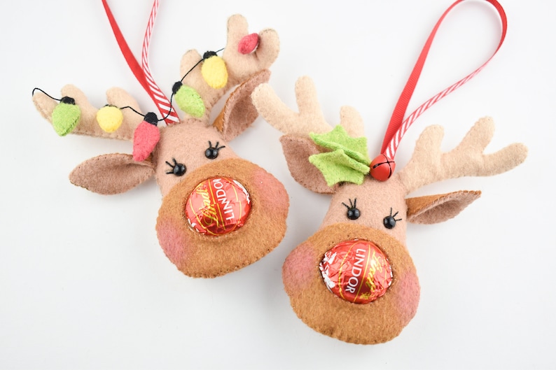 Puede incluir: Dos adornos de reno de fieltro con cintas de suspensi&oacute;n a rayas rojas y blancas. Cada reno tiene una trufa de chocolate Lindor en la boca y una nariz de fieltro. Un reno tiene hojas de acebo de fieltro verde en sus cuernos y el otro tiene una cadena de luces de fieltro amarillo y verde.