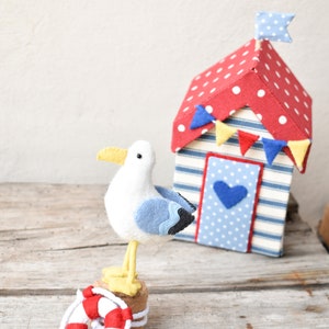 Op de afbeelding: Een witte viltmeeuw met blauwe vleugels staat op een reddingsboei en een houten basis. Achter de meeuw staat een rood-witte geblokte strandhuisje met blauw-witte strepen en een blauw hart.