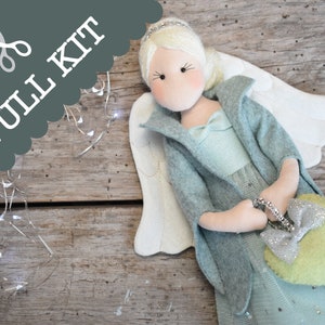 May include: A full kit to make a felt angel doll with a light blue dress, a grey jacket, and white wings. The angel is holding a green heart-shaped purse with silver glitter.