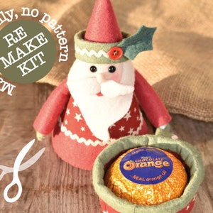 May include: A red and white felt Santa Claus ornament with a green holly sprig on his hat. The ornament is open to reveal a chocolate orange wrapped in gold foil. The text on the foil reads "Terry's Chocolate Orange...REAL orange oil". The image also includes a green circle with the text "RE MAKE KIT Materials only, no pattern".