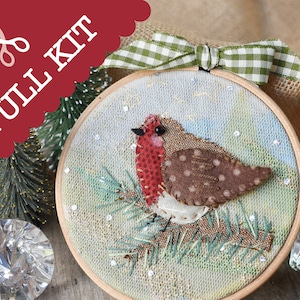 Peut inclure: Un kit complet pour une décoration de Noël en feutre représentant un rouge-gorge perché sur une branche de pin verte sur fond enneigé. L'ornement est encadré dans un cercle en bois avec un ruban vichy vert et blanc.