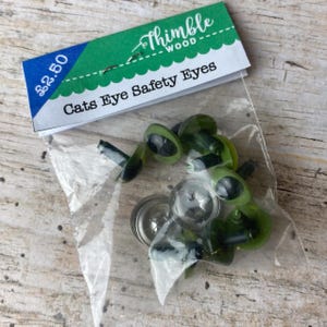 May include: A clear plastic bag containing several green and black cat's eye safety eyes for crafts. The bag has a label with the text "Cats Eye Safety Eyes" and the Thimble Wood logo.