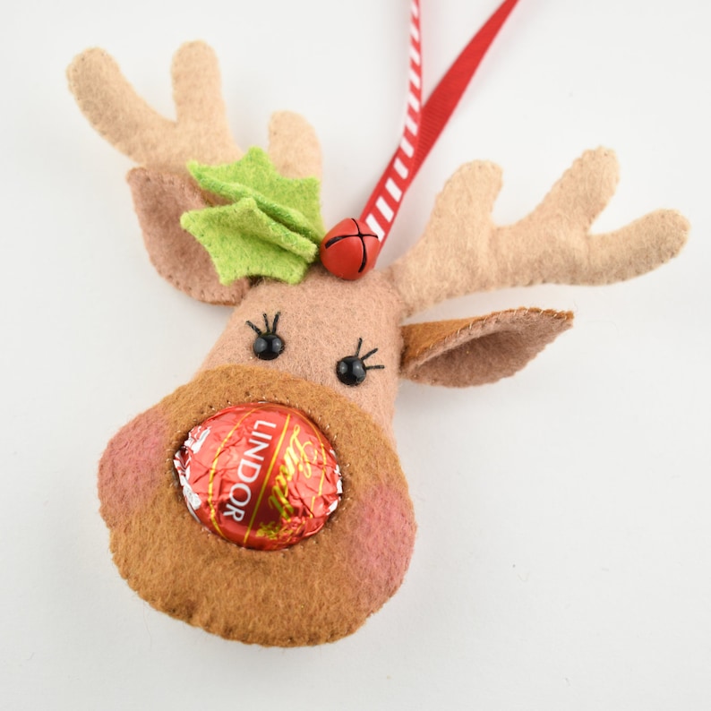 Puede incluir: Un adorno de reno de fieltro con una cinta roja y blanca a rayas, una hoja de acebo verde, una campana roja y una trufa de chocolate Lindor.