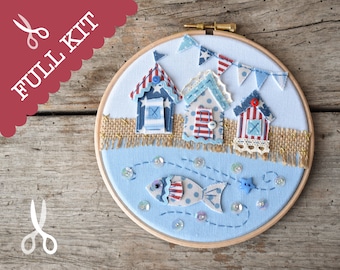 May include: A blue and white fabric hoop art piece depicting three beach houses with red and white striped roofs, a blue and white fish, and a blue star. The houses are on a sandy beach with a burlap border. The hoop is made of wood.
