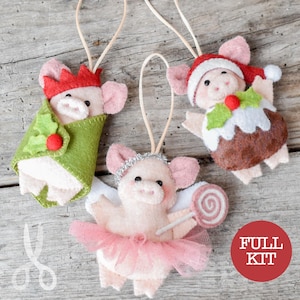 May include: Three felt pig ornaments with hanging loops. One pig wears a red crown and green wrap with holly, one wears a Santa hat and pudding costume, and one wears a silver headband, pink tutu, and holds a pink and white swirl lollipop.