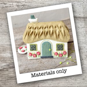 Könnte beinhalten: Ein weißes Filzhaus-Ornament mit einem strukturierten beigen Dach, einer türkisfarbenen Bogentür mit kleinem grünen Knopf, zwei dunkel-türkisfarbenen Fenstern mit Kreuzstich und roten Blumenapplikationen. Der Text "Materials only, no pattern" ist zu sehen.