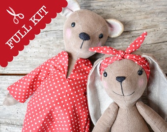 Puede incluir: Dos peluches hechos de tela de pana color canela. Uno lleva un vestido rojo con lunares blancos y el otro una diadema roja con lunares blancos. Ambos tienen rasgos faciales bordados, mejillas rosadas y una etiqueta roja que dice FULL KIT.