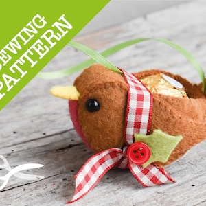 May include: A felt robin ornament with a red and white checkered ribbon, green holly leaves, and a red button. The robin is holding a chocolate treat inside its body.