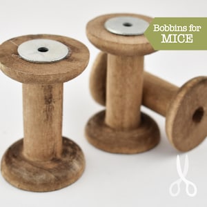 May include: Three wooden spools with metal rings on top. The spools are brown and have a rough texture. The text "Bobbins for MICE" is written on a green background.