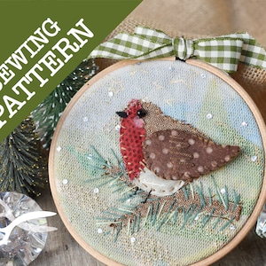 May include: A handmade Christmas ornament featuring a red robin perched on a green pine branch. The ornament is framed in a wooden hoop and decorated with white snowflakes. The background is a light blue and white fabric with a subtle texture.