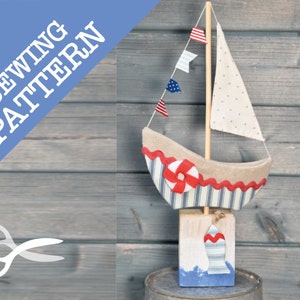 Pode incluir: Um veleiro de tecido feito à mão com detalhes em vermelho, branco e azul. O veleiro está sobre uma base de madeira com um design listrado azul e branco. O veleiro é decorado com tecido listrado vermelho e branco e um salva-vidas vermelho e branco.