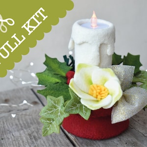 May include: A felt Christmas candle centrepiece with a white candle, a red base, green holly leaves, a white flower with a yellow centre, and a silver ribbon. The text "FULL KIT" is in the top left corner of the image.