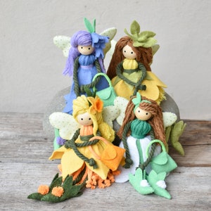 Pode incluir: Quatro bonecas de fada de feltro feitas à mão com asas de flores e carregando pequenas bolsas. As fadas estão vestidas em tons de azul, verde, amarelo e branco.