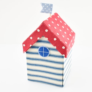 Op de afbeelding: Een klein, rood en wit gestippeld stoffen huis met een blauw en wit gestreept stoffen lichaam en een blauw stoffen raam. Het huis heeft een kleine blauwe en witte vlag bovenop.