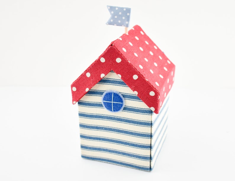 Op de afbeelding: Een klein, rood en wit gestippeld stoffen huis met een blauw en wit gestreept stoffen lichaam en een blauw stoffen raam. Het huis heeft een kleine blauwe en witte vlag bovenop.