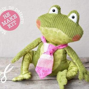 Puede incluir: Un peluche de rana verde con una corbata rosa y blanca estampada. La rana tiene ojos blancos grandes y mejillas rosadas, hecha de pana y tela estampada. La imagen incluye el texto "RE MAKE KIT".
