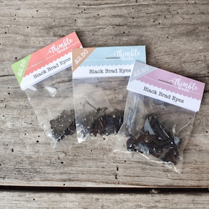 Peut inclure: Trois sachets en plastique transparent contenant des yeux brad noirs, chacun avec une carte d'en-tête étiquetée Thimble Wood et Black Brad Eyes. Les sachets présentent des accents colorés corail, bleu clair et lavande, avec des prix de 2.50.
