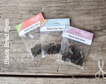 Op de afbeelding: Drie doorzichtige plastic zakjes met zwarte brad-ogen, elk met een kopkaartje met het label Thimble Wood en Black Brad Eyes. De zakjes hebben kleurrijke accenten in koraal, lichtblauw en lavendel, met zichtbare prijskaartjes van 2.50.