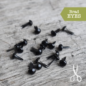 May include: A close-up of black plastic brad eyes, scattered on a wooden surface. The brad eyes are used for crafts and projects.
