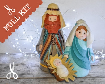 May include: A felt Christmas nativity set with a full kit label. The set includes Joseph, Mary, and baby Jesus. Joseph is wearing a brown robe with a colorful patterned vest. Mary is wearing a blue robe with a white shawl. Baby Jesus is lying in a manger with a yellow halo.