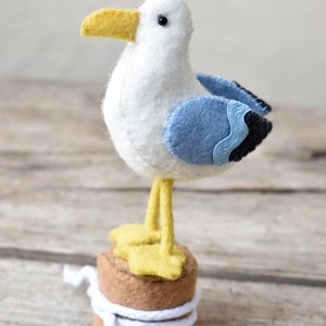 Op de afbeelding: Een witte vilt zeemeeuw met gele snavel en blauwe vleugels staat op een bruine vilt basis met wit touw eromheen gewikkeld.