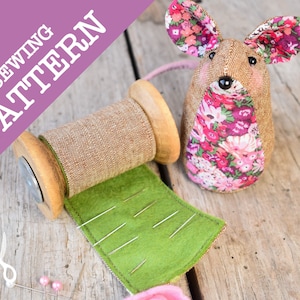 May include: A sewing pattern for a brown and pink floral mouse with a pink tail. The pattern is displayed on a wooden table with a spool of thread, a green felt pincushion, and pink beads.
