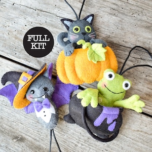 May include: A craft kit featuring three felt ornaments: a grey cat sitting in an orange pumpkin, a grey bat wearing a purple and orange hat with a white bowtie, and a green frog emerging from a black cauldron. Text reads: FULL KIT.