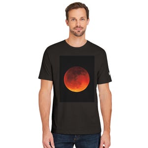 Può includere: Una t-shirt nera a maniche corte con una grande grafica centrale di un'eclissi lunare rossa e arancione su sfondo scuro. La maglietta presenta un piccolo logo bianco sulla manica sinistra ed è indossata con jeans blu scuro.