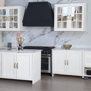 May include: White kitchen cabinets with gold hardware, a black range hood, and a marble countertop. The cabinets have glass doors and drawers.